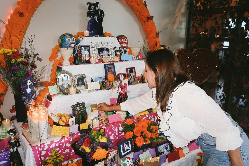 Una mujer se acerca a la ofrenda comunitaria, adornada con fotos enmarcadas de seres queridos, notas escritas a mano, velas, calaveritas y un arco de cempasúchil