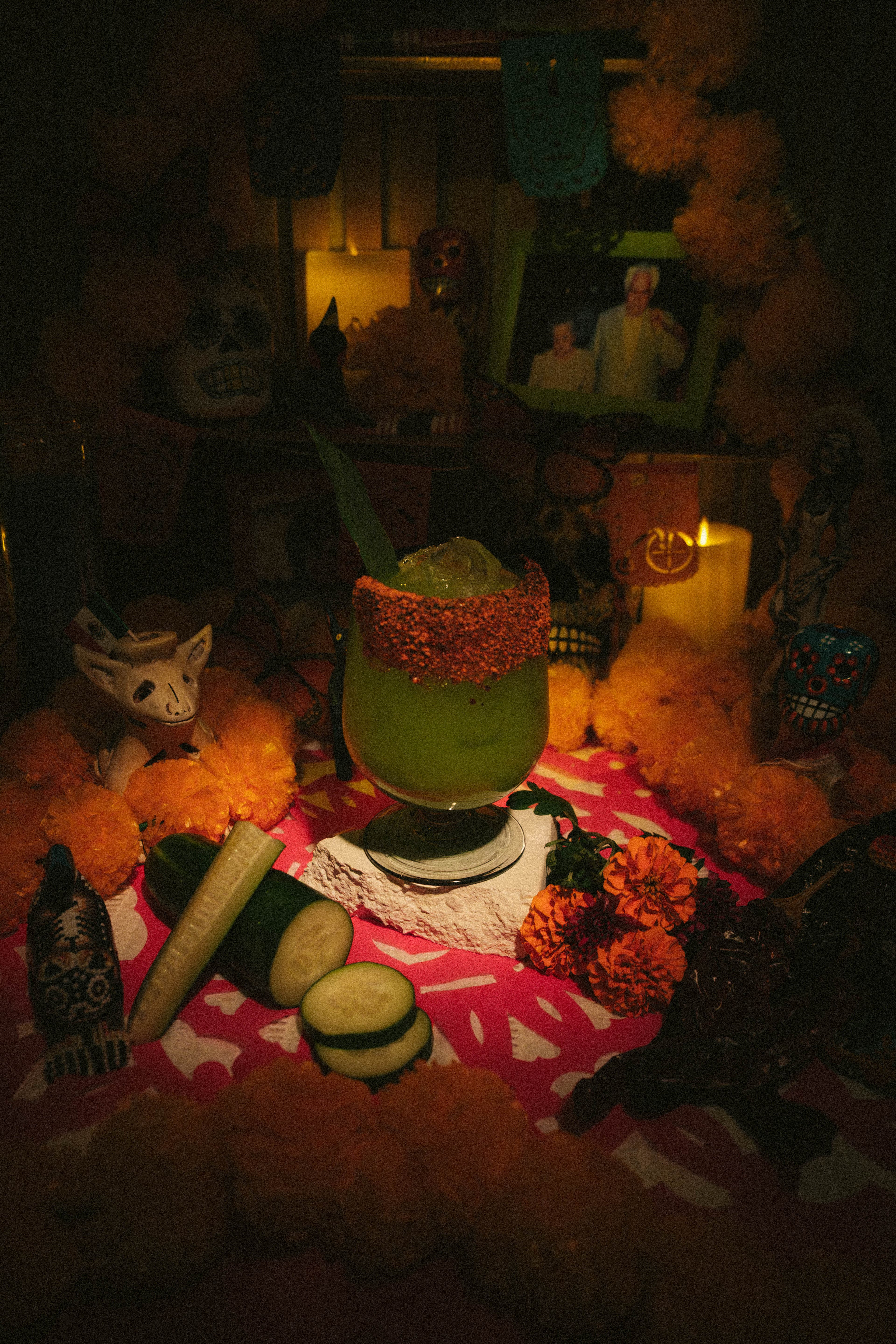 A green agua fresca with a red chamoy and Tajín rim on the ofrenda, beside sliced cucumber and marigolds