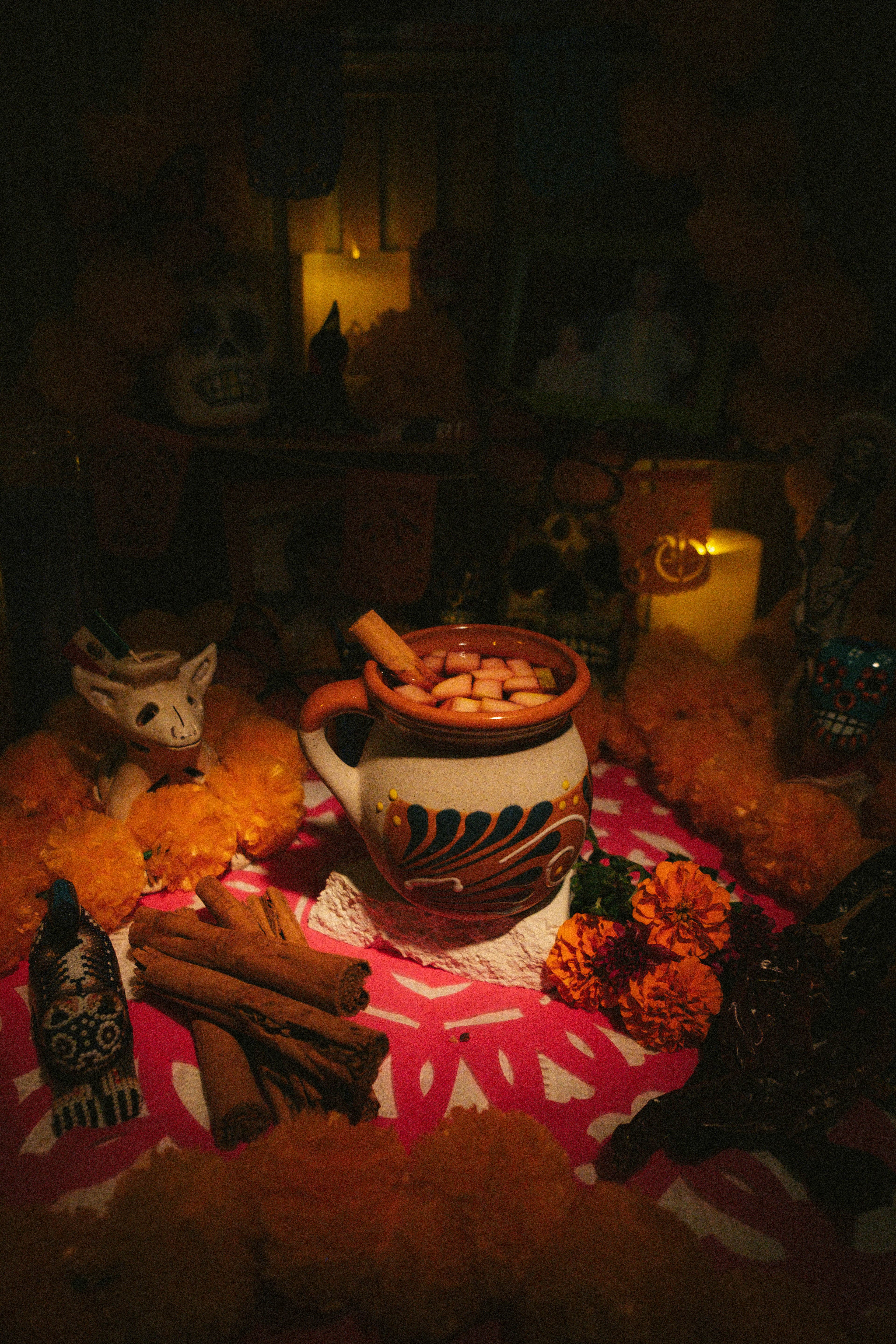 Mexican ponche with diced fruit and a cinnamon stick in a painted clay mug on the ofrenda, beside cinnamon sticks and marigolds