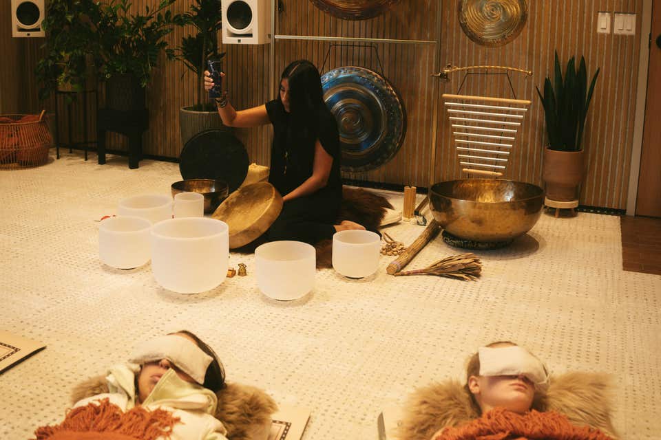 A guide plays a hand drum among crystal bowls and gongs while guests rest under blankets and eye covers at Sacred Society